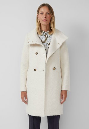 Femme portant un manteau texturé blanc arrivant aux genoux avec de grands revers sur un chemisier floral et un pantalon sombre, se tenant devant un fond gris clair.