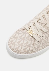 Beige sneaker met een bovenwerk van textiel met een geometrisch patroon, voorzien van een witte zool en een gouden logo op het vetergebied.