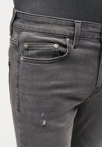 Κοντινή λήψη γκρι τζιν τζινών που φοριούνται με μαύρο τοπ, που δείχνει θηλιές ζώνης, καρφιά και ένα μικρό σκίσιμο στο μηρό.