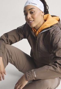 Jeune femme aux cheveux tressés portant un bandeau blanc Terrex, un sweat à capuche orange et une veste et un pantalon Climaproof marron, assise avec un genou levé.