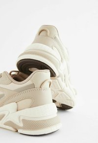 Zapatillas deportivas en color beige con un upper texturizado, que cuentan con una suela más gruesa de goma con patrón y un forro acolchado en el talón.