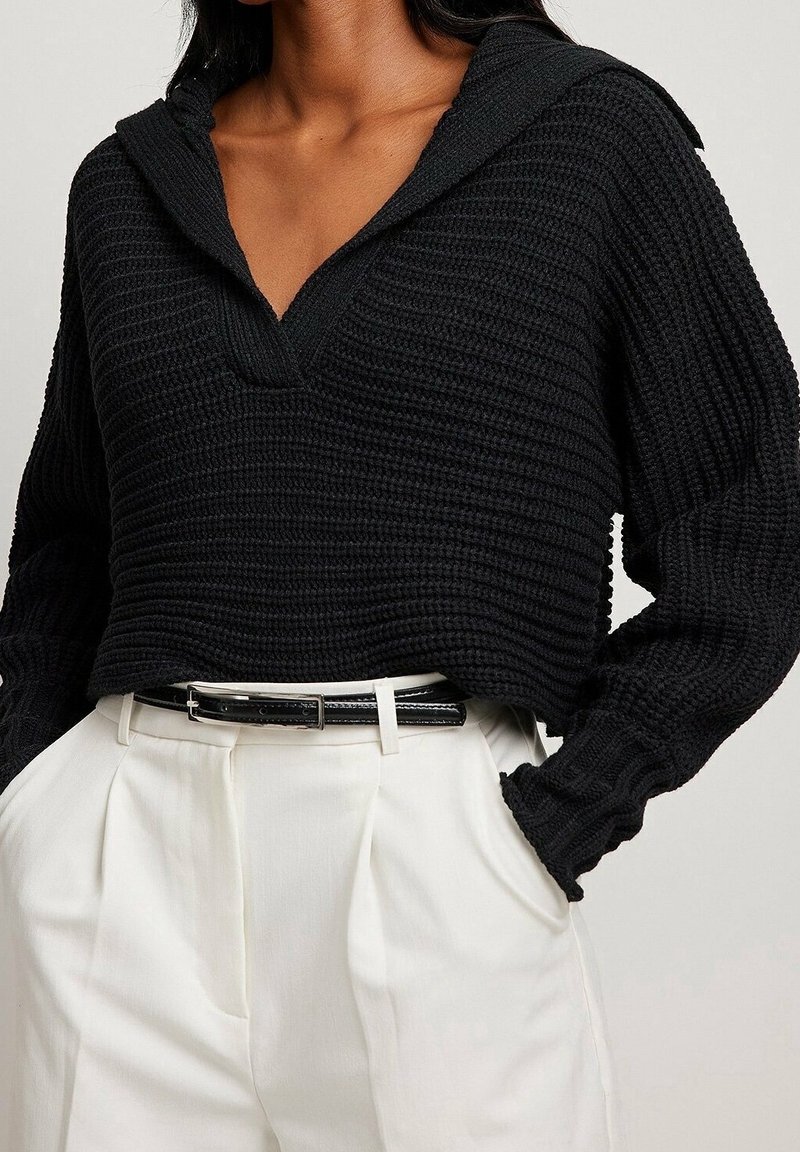 Femme portant un pull à capuche en maille texturée noire, rentré dans un pantalon plissé taille haute blanc avec une ceinture noire.