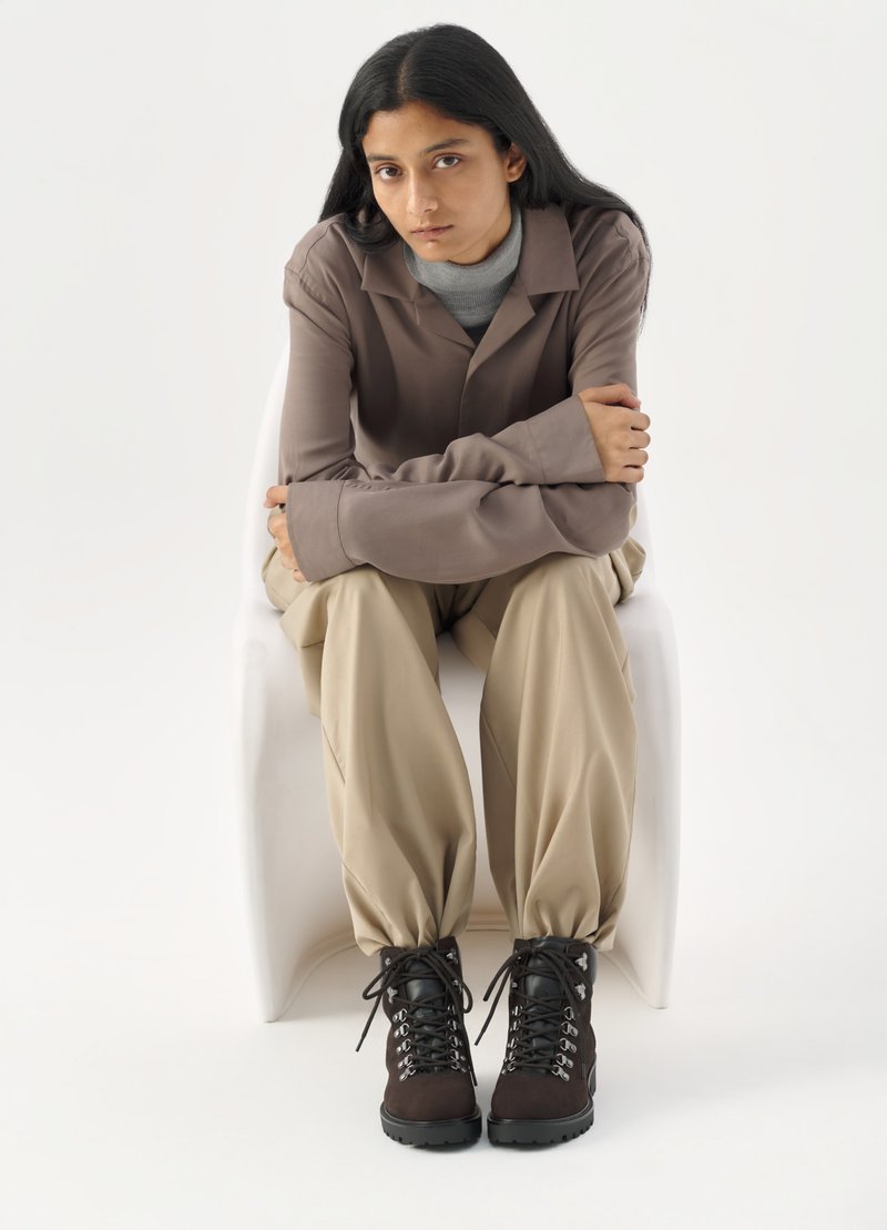 Bruine lange mouwen overhemd over een grijze coltrui, beige losse broek en zwart/bruine veterschoenen met metalen oogjes, zittend op een witte stoel.