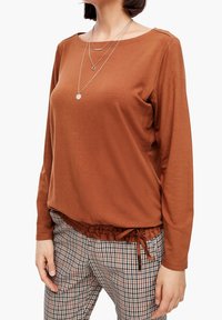 Brązowa bluzka z długim rękawem, okrągłym dekoltem, teksturowanym materiałem i marszczonym wykończeniem na dolnej krawędzi. W połączeniu z spodniami w kratę z ściągaczem.