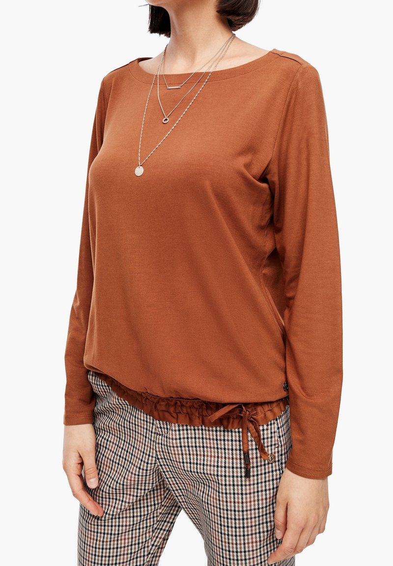 Brązowa bluzka z długim rękawem, okrągłym dekoltem, teksturowanym materiałem i marszczonym wykończeniem na dolnej krawędzi. W połączeniu z spodniami w kratę z ściągaczem.