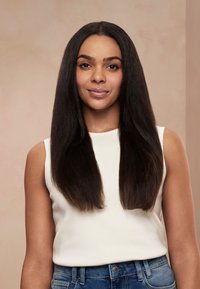 Femme aux longs cheveux lisses et foncés, portant un haut blanc sans manches et un jean bleu, debout devant un fond beige.