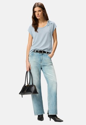 Mujer con top azul claro con nudo en el hombro, vaqueros de mezclilla claros con aberturas en los bajos, cinturón negro, botas negras de tacón y llevando un bolso negro.