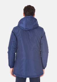 Uomo che indossa una giacca invernale con cappuccio blu navy vista di spalle su uno sfondo bianco semplice.