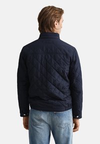 Temno modra prešita jakna z zapenjanjem na pritisne gumbe na manšetah in visokim ovratnikom. Ima gladko teksturo in logotipni obliž na hrbtni strani.