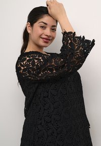 Sort blonde-top med lange, udvidede ærmer, der har et blomsterdesign. Stoffet har et struktureret, delikat udseende.