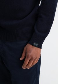 Marineblauer Pullover aus weichem, strukturiertem Material mit geripptem Saum und Bündchen. Verfügt über ein kleines schwarzes Etikett mit weißem Text auf dem Bündchen.