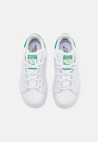 White leather sneakers with green accents and branding, featuring flat laces, a smooth texture, and a low-profile silhouette.