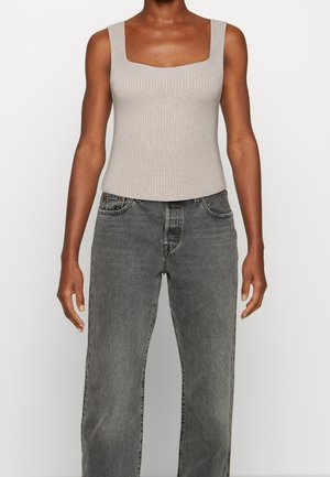 Persona che indossa un top smanicato beige a coste e jeans grigi larghi a vita alta, in piedi davanti a uno sfondo bianco uniforme.