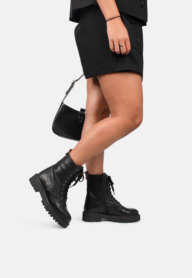 Botas de cuero negras hasta el tobillo con un panel lateral texturizado, cordones en la parte delantera, cremallera lateral y suela resistente. Llevadas con una falda negra.
