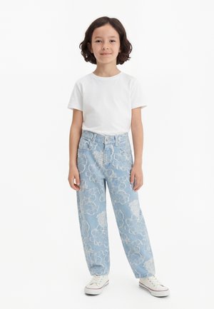 Dieťa s krátkymi kučeravými vlasmi, ktoré má na sebe biele tričko, svetlomodré rifle s kvetinovým vzorom a biele tenisky, stojace proti jednoliatemu pozadiu.