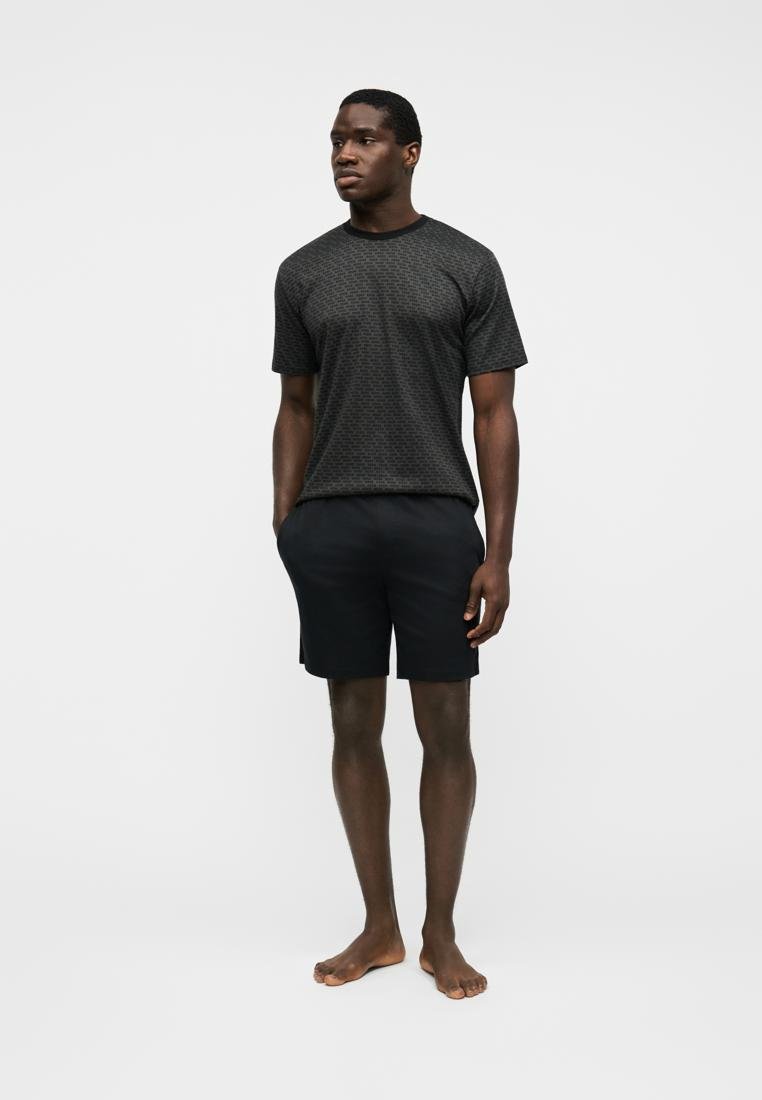 Homme debout pieds nus portant une chemise sombre à manches courtes avec motif et un short noir uni sur un fond blanc uni.