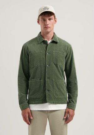 Green corduroy jacket with a button-up front, two front pockets, and a collared neck. Worn over a white shirt.