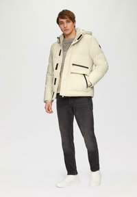 Beige Pufferjacke mit schwarzen Reißverschlussakzenten und vier Taschen, getragen über einem grauen Pullover und kombiniert mit dunkelblauen Jeans und weißen Sneakers.