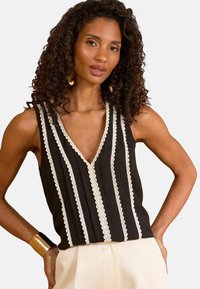 Top sans manches noir avec un col en V, orné de bordures en dentelle blanche verticales et d'un design de tissu texturé, associé à un pantalon de couleur crème.