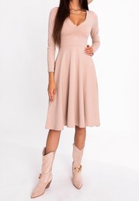 Robe longue rose clair à manches longues avec un col en V et un détail froncé au niveau du bustier, jupe évasée, associée à des bottines beige avec un accent doré.