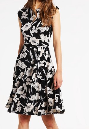 Femme portant une robe noire longueur genou avec des motifs floraux blancs et une ceinture à la taille, debout devant un fond blanc.