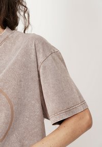 Jasnobrązowa, krótka koszulka z krótkim rękawem, wykonana z miękkiej tkaniny. Posiada okrągły dekolt oraz prosty wzór graficzny w ciemniejszym odcieniu.