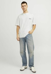 Hvid kortærmet t-shirt med et sort logo på venstre bryst, parret med lyseblå lige ben jeans og hvide sneakers med sorte accenter.