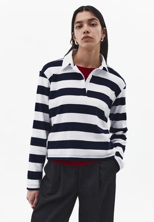 Kobieta o długich ciemnych włosach nosi srebrne kolczyki koła, granatowo-biały pasiasty longsleeve, czerwoną podkoszulkę i ciemne spodnie w cienkie prążki.