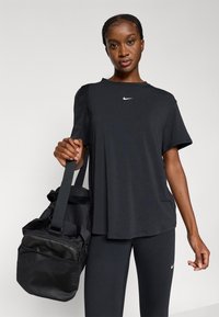 Juoda Nike marškinėliai su apvalia iškirpte ir trumpomis rankovėmis. Derinami su juodomis tampriomis kelnėmis ir laikomas juodas sportinis krepšys su tekstūruotu paviršiumi.