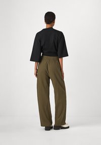 Czarna, oversize koszula z szerokimi rękawami, zestawiona z luźnymi plisowanymi spodniami w oliwkowym kolorze. Model stoi tyłem do kamery, mając na nogach białe buty na platformie.