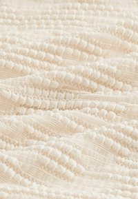 Tissu crème texturé présentant des côtes et des boucles en relief, avec une apparence douce et réconfortante. La surface affiche un motif ondulé subtil.