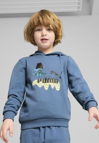 Modrá mikina s kapucňou má na prednej strane grafiku modrej animovanej postavy a logo "PUMA". Mäkko tkanina, ribbované manžety a lemovanie.