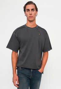 Ung mann med bakoverkjemmet hår iført en mørkegrå overdreven T-skjorte og blå jeans, stående mot en enkel hvit bakgrunn.