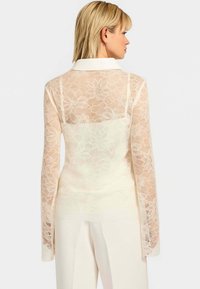 Blondebluse i off-white med blomsterprint, lange ærmer og krave. Bagstykket er semitransparent og lagdelt over en solid bund.