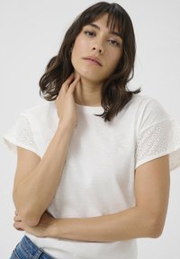 T-shirt blanc à manches courtes avec des manches en dentelle brodée. Tissu doux et texturé avec une coupe décontractée et un col rond.