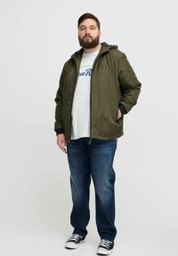 Jachetă capucioasă de culoare verde măsliniu, cu fermoar frontal, cu manșete negre și buzunare, purtată peste un tricou alb cu grafică și blugi, completată cu adidași.
