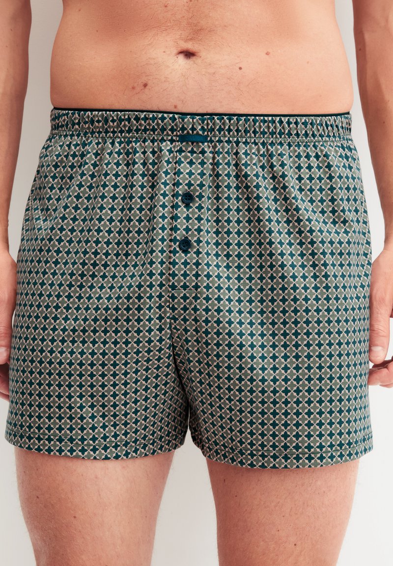 Boxershorts mit einem geometrischen Muster in Türkis und Beige, ausgestattet mit einem Knopfverschluss und einem elastischen Bund für Komfort. Glatte Stoffstruktur.
