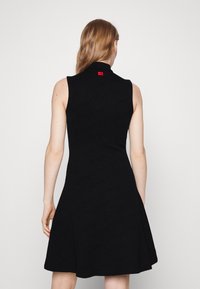 Robe noire sans manches avec une silhouette évasée, un matériau texturé et une étiquette rouge au col arrière.