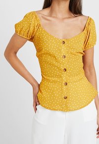 Blusa gialla a pois con maniche a sbuffo, scollatura a barco e bottoni frontali. Realizzata in tessuto strutturato con un design aderente.