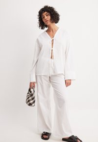 Valge linane kostüüm, mis sisaldab vabalt langevat pluusi, millel on kaela juures nöörid, ja laiad püksid. Must tekstureeritud käekott ja sandaalid täiustavad välimust.