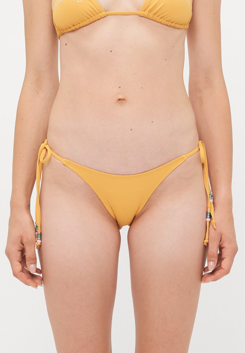 Frau, die einen gelben Bikini mit Seitenbändern und Perlenbindern sowie einem passenden Bikinioberteil trägt, steht vor einem schlichten Hintergrund.