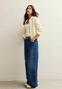 Crème gebreide cardigan met een kraag, gedragen over een witte tanktop. Gecombineerd met hoge, wijde blauwe jeans en witte schoeisel.
