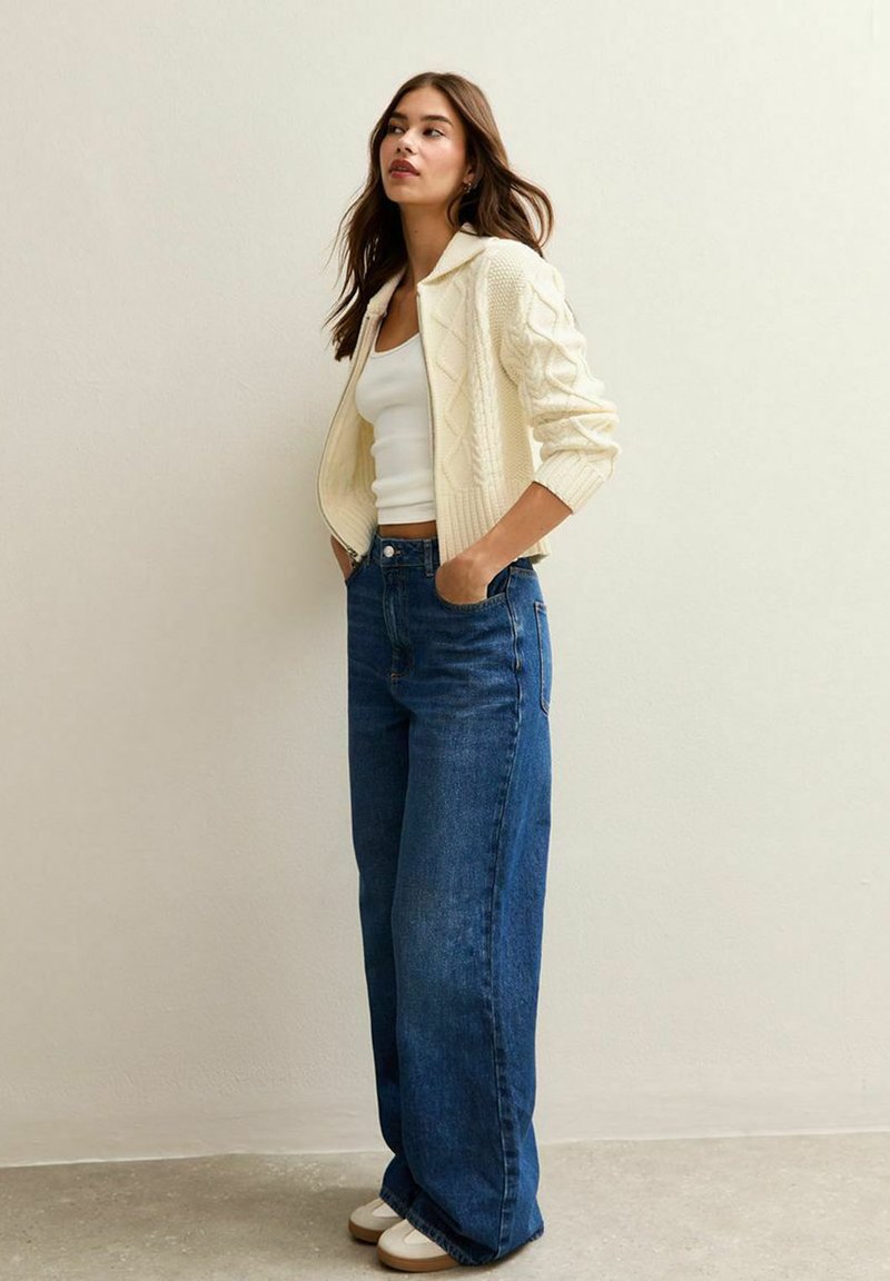 Crème gebreide cardigan met een kraag, gedragen over een witte tanktop. Gecombineerd met hoge, wijde blauwe jeans en witte schoeisel.