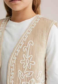 Gilet beige ricamato con motivi floreali bianchi e dettagli texture, indossato sopra una camicia bianca a maniche lunghe.