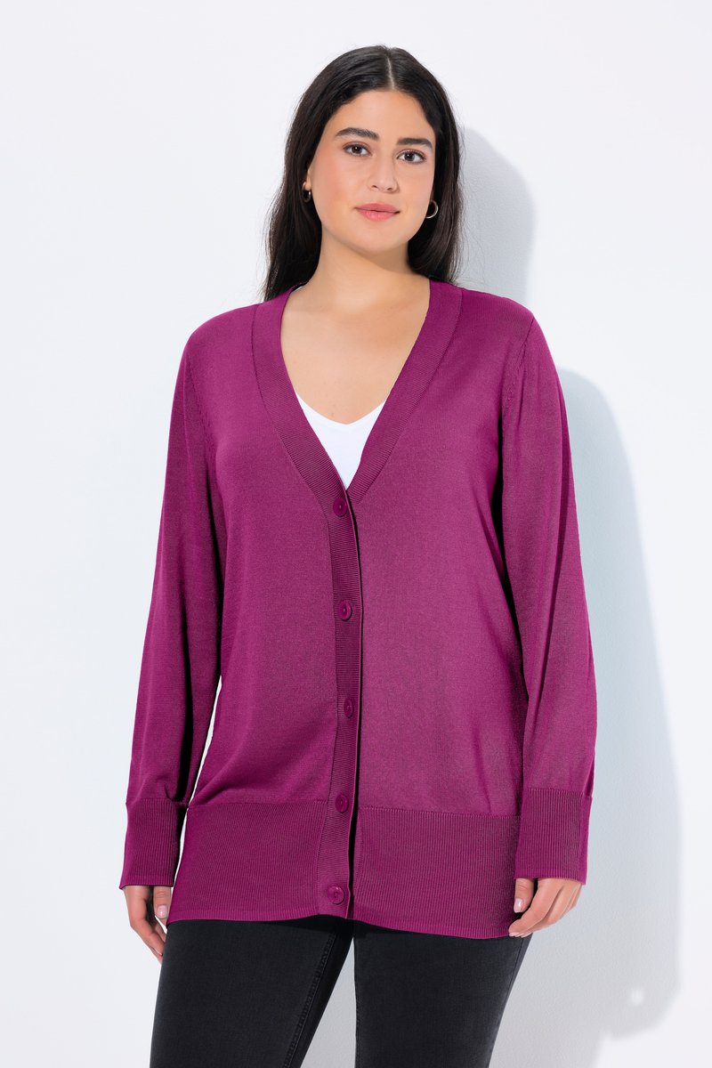 Frau mit dunklem Haar trägt eine lila geknöpfte Strickjacke über einem weißen Top und schwarzen Hosen, steht vor einem schlichten hellen Hintergrund.