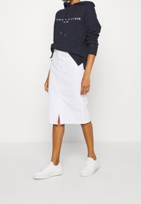 Personne portant un hoodie navy Tommy Hilfiger, une jupe blanche mi-longue avec une fente à l'avant, et des baskets blanches, se tenant devant un fond uni.