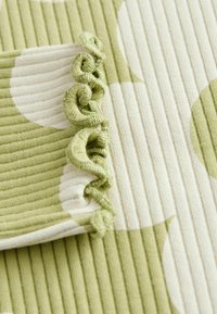 Grün- und cremefarbenes Gewebe mit gerippter Textur. Auffällige Rüschendetails am Rand, mit großen kreisförmigen Mustern in verschiedenen Grüntönen.