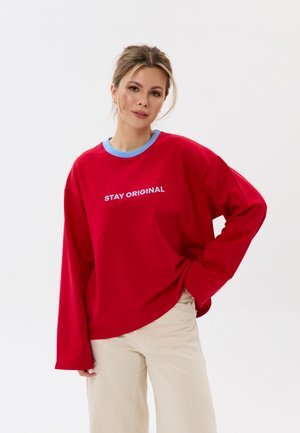 Femme aux cheveux clairs portant une chemise rouge à manches longues avec un col bleu et le texte "STAY ORIGINAL", associée à un pantalon beige, posant devant un fond blanc.