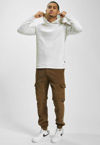 Hvit hettegenser med høy halsdesign, parret med brune corduroy cargo-bukser med sidelommer, og hvite joggesko med svarte accenter.