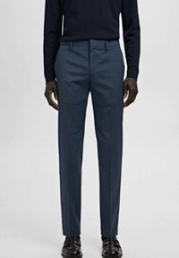 Mężczyzna ubrany w dopasowane granatowe spodnie garniturowe, czarną bluzkę z długim rękawem oraz czarne skórzane eleganckie buty, stojący na jednolitym tle.